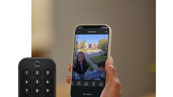 Photo of a person’s hand holding a phone running the Ecobee app. It shows a live view of two children at the door. A Yale smart lock is overlaid to the lower left.